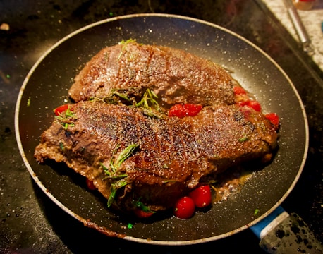 Two richly-browned steaks seasoned with herbs are sizzling in a pan atop a bed of cherry tomatoes. The surface of the meat is textured and glistening, with sprigs of rosemary adding a fresh, aromatic touch. The scene is set on a dark stovetop, creating a warm, appetizing appearance.