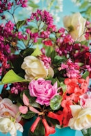A vibrant floral arrangement featuring a variety of flowers in shades of pink, red, and cream. The composition includes roses, delicate clusters of smaller blossoms, and lush green leaves, all set against a soft blue background.
