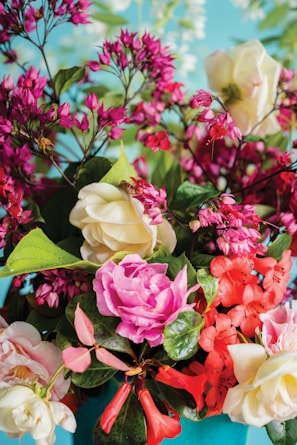 A vibrant floral arrangement featuring a variety of flowers in shades of pink, red, and cream. The composition includes roses, delicate clusters of smaller blossoms, and lush green leaves, all set against a soft blue background.