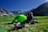 A person is seated on a portable chair in front of a bright green tent situated on a grassy plain. In the background are majestic mountains with rocky peaks and some snow patches under a clear blue sky. The individual appears to be enjoying a snack, surrounded by camping gear and a red bottle.