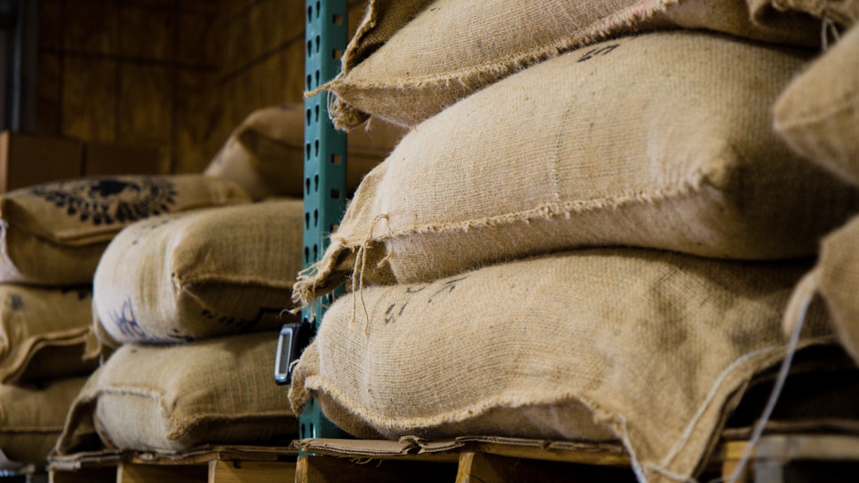 Stacks of burlap sacks resting on wooden pallets are arranged in a warehouse or storage facility. The sacks, likely filled with agricultural products like coffee beans or grains, appear weathered yet sturdy. The industrial shelving provides support to the weight of the sacks.
