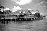 A large house with intricate detailing is built on stilts over the water, featuring a sloping roof and multiple windows. In the background, a tall, modern building and lush trees create a contrast with the traditional architecture. The sky is partly cloudy, adding texture to the scene.