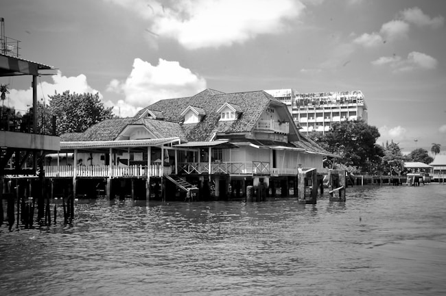 A large house with intricate detailing is built on stilts over the water, featuring a sloping roof and multiple windows. In the background, a tall, modern building and lush trees create a contrast with the traditional architecture. The sky is partly cloudy, adding texture to the scene.