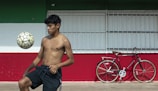 A shirtless young man balances a soccer ball on his knee in an outdoor setting with a large horizontal tricolor wall of green, white, and red. To the right, against the wall, is a parked black bicycle with a brown seat and handlebars.