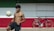 A shirtless young man balances a soccer ball on his knee in an outdoor setting with a large horizontal tricolor wall of green, white, and red. To the right, against the wall, is a parked black bicycle with a brown seat and handlebars.