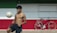 A shirtless young man balances a soccer ball on his knee in an outdoor setting with a large horizontal tricolor wall of green, white, and red. To the right, against the wall, is a parked black bicycle with a brown seat and handlebars.