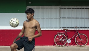 A shirtless young man balances a soccer ball on his knee in an outdoor setting with a large horizontal tricolor wall of green, white, and red. To the right, against the wall, is a parked black bicycle with a brown seat and handlebars.