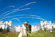 A couple in wedding attire embraces in front of a large, white castle with turrets under a clear blue sky. Tall grasses sway in the foreground, capturing a sense of openness and tranquility.