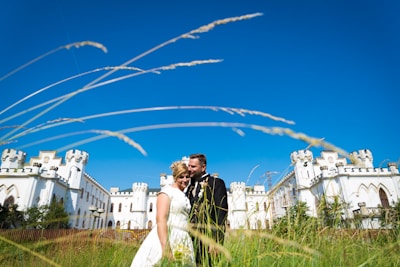 A couple in wedding attire embraces in front of a large, white castle with turrets under a clear blue sky. Tall grasses sway in the foreground, capturing a sense of openness and tranquility.