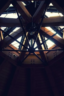 A structural ceiling features a geometric pattern with wooden beams and dark metal rods forming an intricate design. Natural light filters through the open sections, creating contrast between the illuminated and shaded areas.