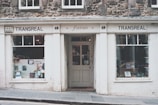 A quaint bookstore with a vintage façade named Transreal, showcasing a variety of books in its display windows. The building has stonework and a classic design, with large windows on either side of a central door. The signage includes the word 'fiction', indicating a focus on literary works.