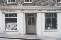 A quaint bookstore with a vintage façade named Transreal, showcasing a variety of books in its display windows. The building has stonework and a classic design, with large windows on either side of a central door. The signage includes the word 'fiction', indicating a focus on literary works.