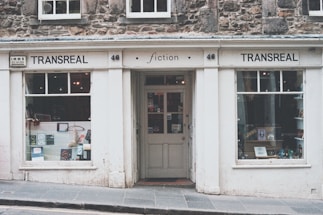 A quaint bookstore with a vintage façade named Transreal, showcasing a variety of books in its display windows. The building has stonework and a classic design, with large windows on either side of a central door. The signage includes the word 'fiction', indicating a focus on literary works.