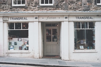 A quaint bookstore with a vintage façade named Transreal, showcasing a variety of books in its display windows. The building has stonework and a classic design, with large windows on either side of a central door. The signage includes the word 'fiction', indicating a focus on literary works.