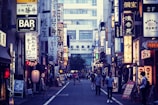 A bustling urban street in Japan with numerous illuminated signs written in Japanese characters lining both sides. The shops and buildings create a vibrant cityscape. People walk along the narrow road, adding a sense of liveliness to the scene.