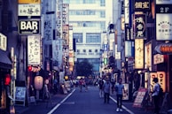 A bustling urban street in Japan with numerous illuminated signs written in Japanese characters lining both sides. The shops and buildings create a vibrant cityscape. People walk along the narrow road, adding a sense of liveliness to the scene.