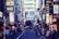 A bustling urban street in Japan with numerous illuminated signs written in Japanese characters lining both sides. The shops and buildings create a vibrant cityscape. People walk along the narrow road, adding a sense of liveliness to the scene.
