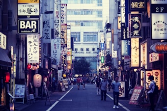 A bustling urban street in Japan with numerous illuminated signs written in Japanese characters lining both sides. The shops and buildings create a vibrant cityscape. People walk along the narrow road, adding a sense of liveliness to the scene.