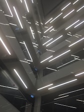 An architectural design featuring multiple levels of staircases and landings, illuminated by bright linear lights. The composition creates geometric patterns and lines, with glass and metal surfaces reflecting light to add depth.