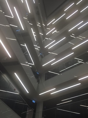 An architectural design featuring multiple levels of staircases and landings, illuminated by bright linear lights. The composition creates geometric patterns and lines, with glass and metal surfaces reflecting light to add depth.