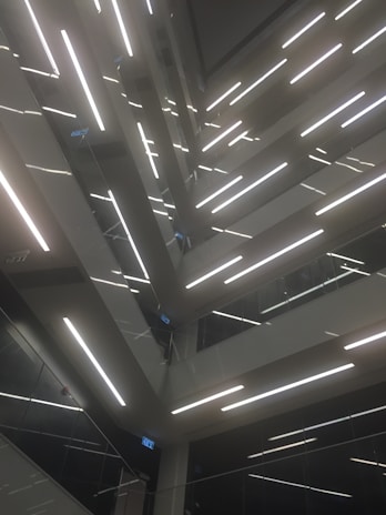 An architectural design featuring multiple levels of staircases and landings, illuminated by bright linear lights. The composition creates geometric patterns and lines, with glass and metal surfaces reflecting light to add depth.