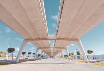 An overhead perspective of a concrete bridge structure supported by tall pillars, with a clear blue sky in the background. The bridge runs parallel to a road below, lined with a few evenly spaced trees. Modern street lamps are visible, and the surroundings hint at an urban setting.
