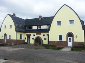 A large house with a unique architectural design featuring two gable-roofed sections connected by a central portion. The building has a soft yellow exterior with dark roofing tiles and red brick accents at the base. Windows are symmetrically arranged across the facade, and there are two prominent entrances, each arched and framed with bricks. The area in front of the house is paved, with some small plants and outdoor furniture, creating a welcoming entrance.