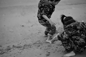 Two children wearing patterned jackets are playing on the sand at a beach. One child is crouching down, possibly building or drawing in the sand, while the other is standing nearby. Their clothing features a camouflage pattern, and they appear engaged and focused on their activity.