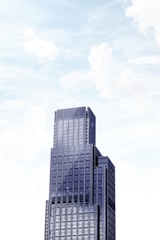 A towering modern skyscraper with multiple tiers against a bright sky filled with soft clouds. The building features a sleek facade with large windows and a geometric design.