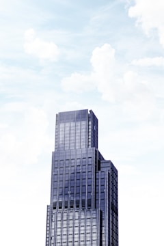 A towering modern skyscraper with multiple tiers against a bright sky filled with soft clouds. The building features a sleek facade with large windows and a geometric design.