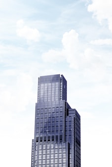 A towering modern skyscraper with multiple tiers against a bright sky filled with soft clouds. The building features a sleek facade with large windows and a geometric design.