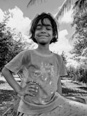 A young child with a confident smile stands outdoors, surrounded by trees and a partly cloudy sky. The child is wearing a T-shirt with the phrase 'I believe I can fly' and illustrations of airplanes.