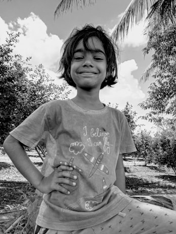 A young child with a confident smile stands outdoors, surrounded by trees and a partly cloudy sky. The child is wearing a T-shirt with the phrase 'I believe I can fly' and illustrations of airplanes.