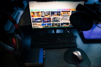 A dimly lit computer workstation featuring a large monitor displaying various colorful images of workspaces. A keyboard and a mouse are positioned on the dark wooden desk. A microphone with a pop filter is visible on the right side, with headphones resting on the left. A small audio interface device is also present beside the keyboard.