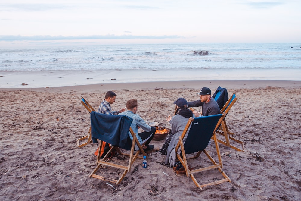 three men and one woman sitting on beach lounge in front fire pit near seashore
