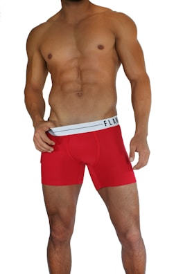 A muscular male torso is displayed, wearing bright red boxer briefs with a white waistband. The waistline of the briefs has a brand logo visible on it. The skin tone is tanned and the individual is posed in a confident stance.