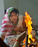 A woman in traditional attire sits near a small fire, appearing to be involved in a ritual. Her saree is adorned with colorful patterns, and she is wearing bangles. The flames create a warm and vibrant contrast against the subdued background.