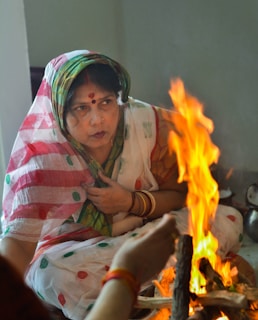 A woman in traditional attire sits near a small fire, appearing to be involved in a ritual. Her saree is adorned with colorful patterns, and she is wearing bangles. The flames create a warm and vibrant contrast against the subdued background.