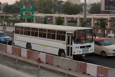 A white bus is traveling on a busy road next to other vehicles, including a blue car and a red car. The scene is set in an urban environment with greenery and road signs indicating directions to different airline terminals. The foreground includes traffic barriers and a construction cone.