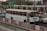 A white bus is traveling on a busy road next to other vehicles, including a blue car and a red car. The scene is set in an urban environment with greenery and road signs indicating directions to different airline terminals. The foreground includes traffic barriers and a construction cone.