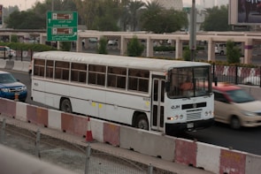 A white bus is traveling on a busy road next to other vehicles, including a blue car and a red car. The scene is set in an urban environment with greenery and road signs indicating directions to different airline terminals. The foreground includes traffic barriers and a construction cone.