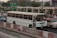 A white bus is traveling on a busy road next to other vehicles, including a blue car and a red car. The scene is set in an urban environment with greenery and road signs indicating directions to different airline terminals. The foreground includes traffic barriers and a construction cone.