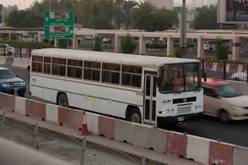 A white bus is traveling on a busy road next to other vehicles, including a blue car and a red car. The scene is set in an urban environment with greenery and road signs indicating directions to different airline terminals. The foreground includes traffic barriers and a construction cone.