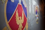 A close-up view of a colorful emblem featuring a stylized golden eagle on a red and blue shield set against a gray background. To the right, there are additional emblems, including a star design, partially visible.
