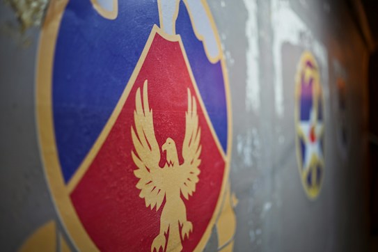 A close-up view of a colorful emblem featuring a stylized golden eagle on a red and blue shield set against a gray background. To the right, there are additional emblems, including a star design, partially visible.