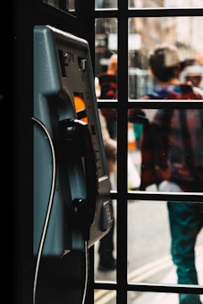 A payphone is mounted inside a booth with a glass window. The foreground features the side view of the phone, including the handset and cord. The background is slightly blurred, showing people walking outside, suggestive of a busy street scene.