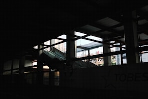 A dimly lit interior space of a construction or industrial site, with visible steel beams and a partially finished structure. Light pours in through large open spaces, casting shadows across the area. An escalator leads upwards amidst the framework.