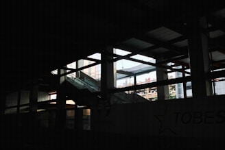 A dimly lit interior space of a construction or industrial site, with visible steel beams and a partially finished structure. Light pours in through large open spaces, casting shadows across the area. An escalator leads upwards amidst the framework.