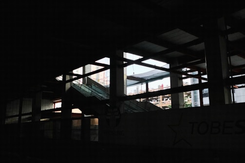 A dimly lit interior space of a construction or industrial site, with visible steel beams and a partially finished structure. Light pours in through large open spaces, casting shadows across the area. An escalator leads upwards amidst the framework.