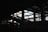 A dimly lit interior space of a construction or industrial site, with visible steel beams and a partially finished structure. Light pours in through large open spaces, casting shadows across the area. An escalator leads upwards amidst the framework.